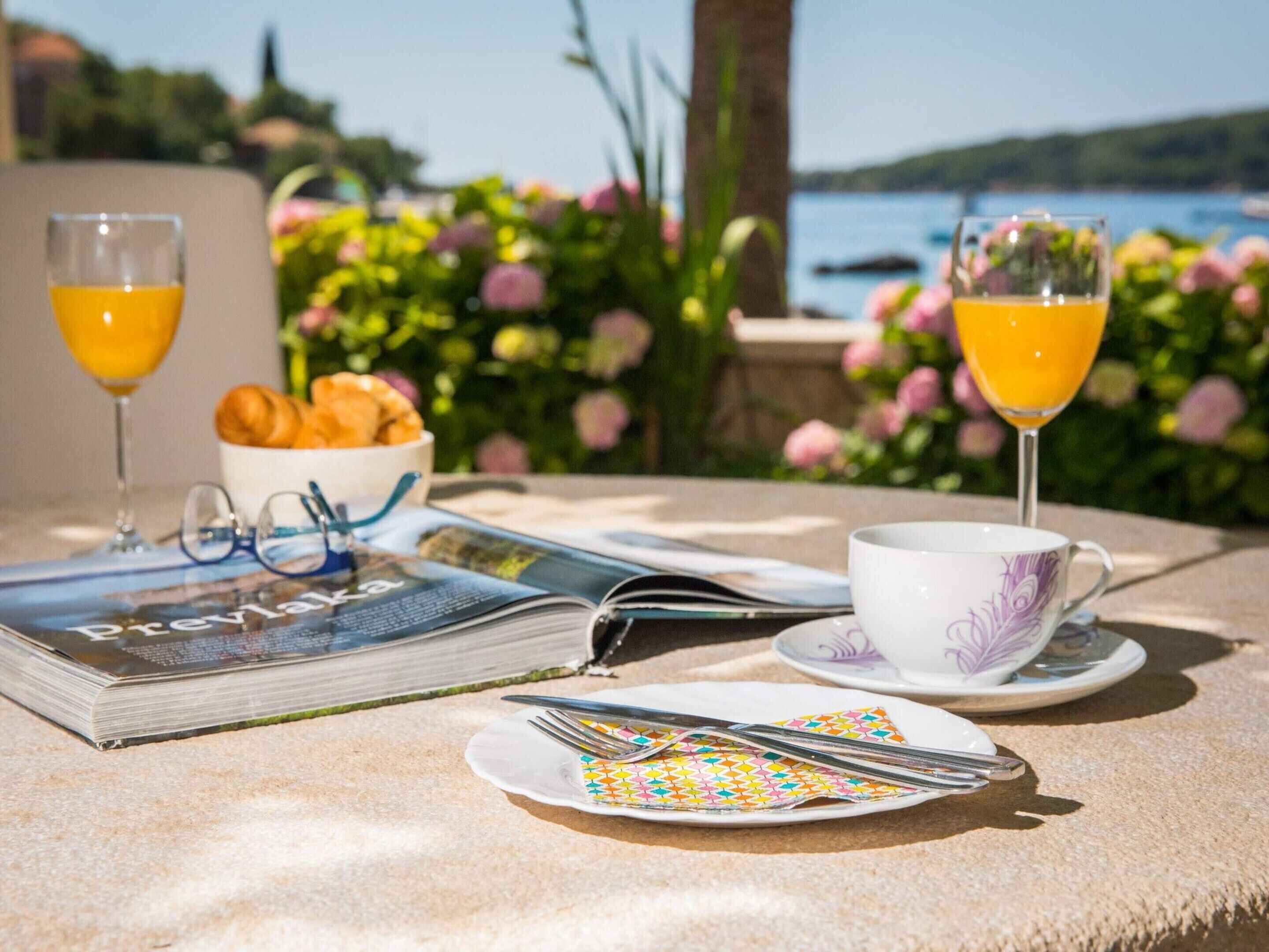 Une table dressée pour le petit-déjeuner, avec des croissants, du jus d’orange, une tasse, un magazine, des verres et des couverts.