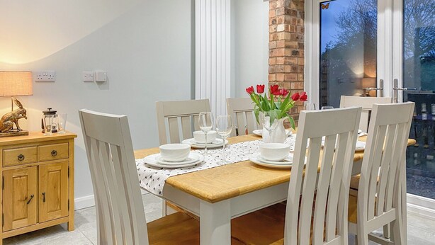 A dining area with a wooden table set for four, white chairs, a vase with red flowers, and a brick wall. - 3 bedroom accommodation in Rugby (Rugby)
