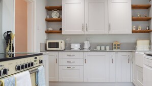 A modern kitchen with white cabinets, a microwave, a kettle, and a toaster. There are wooden shelves above the counter and a clock on the wall. - 3 bedroom accommodation in Rugby (Rugby)