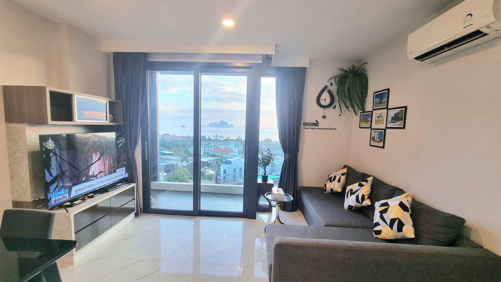 A modern living room with a flat-screen TV, a sofa with patterned cushions, a small side table, and a large sliding glass door leading to a balcony with a city view.