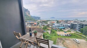 A balcony with a table, chairs, a bottle of wine, and a glass. The view includes a coastal area with buildings, a parking lot, and a mountainous landscape. - Bo501 - 2 Bdr Condo With Sea View, Ao Nang Beach (Krabi)