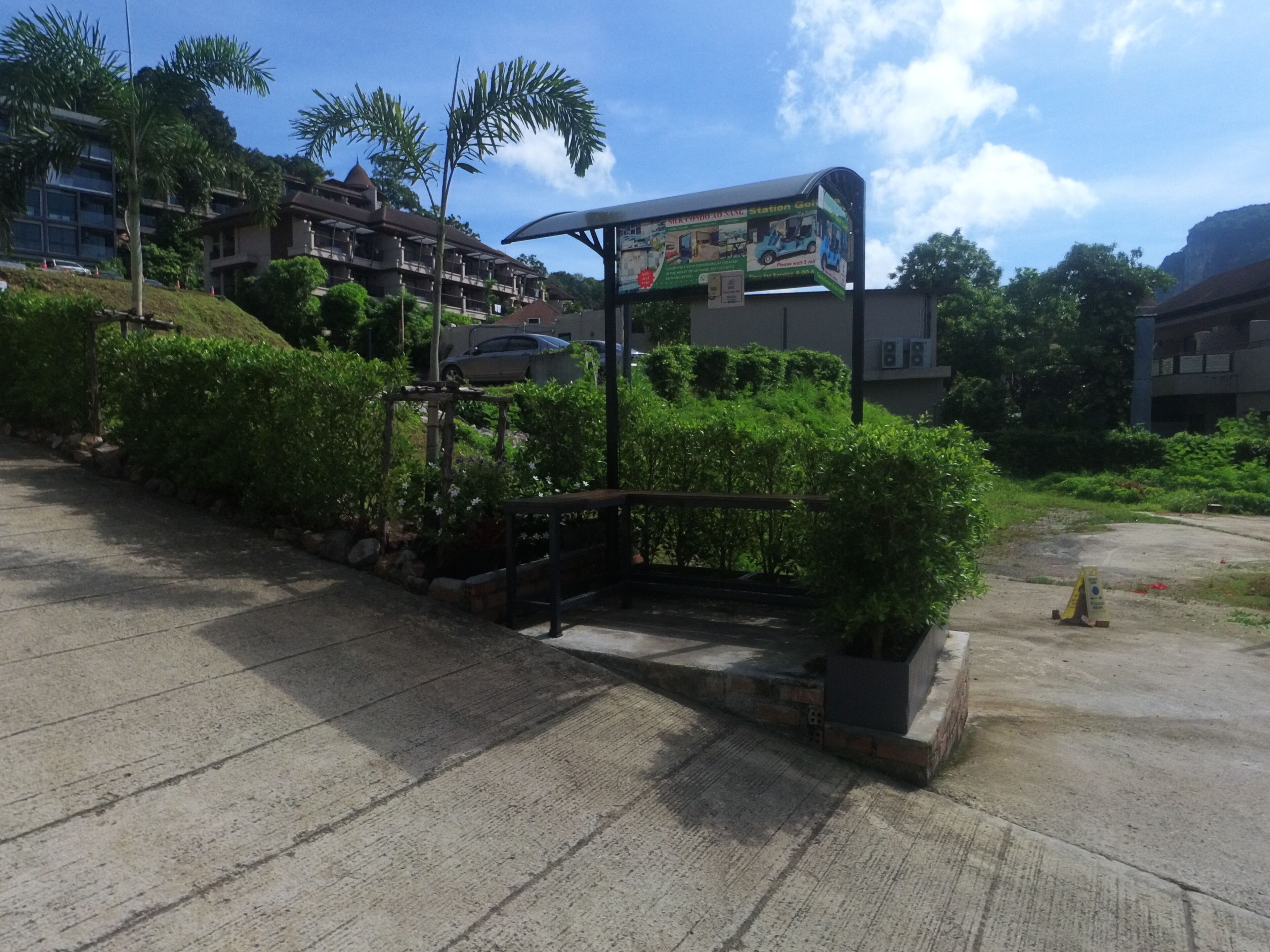 A parking area with a signboard, greenery, and buildings in the background.