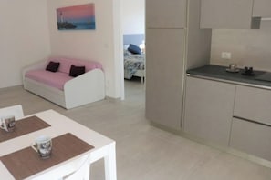 Ein modernes Zimmer mit einer Küchenzeile, einer Essecke mit Tisch und Stühlen, einem rosa Sofa und einem Gemälde eines Leuchtturms.