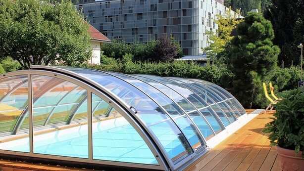 A glass-enclosed swimming pool with a wooden deck, surrounded by greenery and a building in the background. - Apartment Near the Gondelbahn With Shared Pool (Hermagor-Pressegger See)