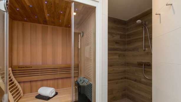 A sauna with a wooden bench and a towel, and a shower area with a wooden wall and a showerhead. - House in Oostkapelle Near Beach & Sauna (Oostkapelle)