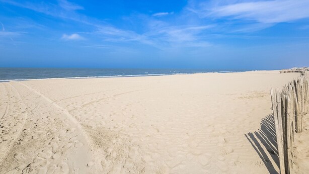 Une vaste plage de sable, avec une clôture en bois sur la droite et un ciel bleu dégagé.