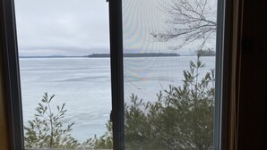 A window with a view of a calm body of water, a distant shoreline, and some trees in the foreground.