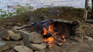 A campfire with rocks around it, a wooden structure, and a view of a lake with trees in the background.