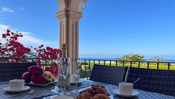 Una mesa dispuesta con una taza blanca, un plato de croissants, un vaso de agua y una botella de champán, con vista al océano y follaje en el fondo.