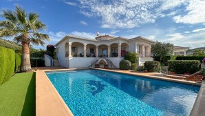 Una piscina con superficie de agua cristalina y azul, rodeada por un césped cuidado y una casa grande con tejado de baldosas y varias ventanas.