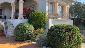 Una casa de dos pisos con balcón, escaleras y un jardín con flores rojas.