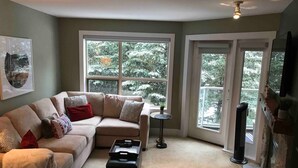 A living room with a beige sectional sofa, a small round table, and a black ottoman. There are large windows with a view of trees and a fireplace. - Slope-side condo ski in ski out! Quiet side of resort, heated pool + 3 hot tubs (Whistler)