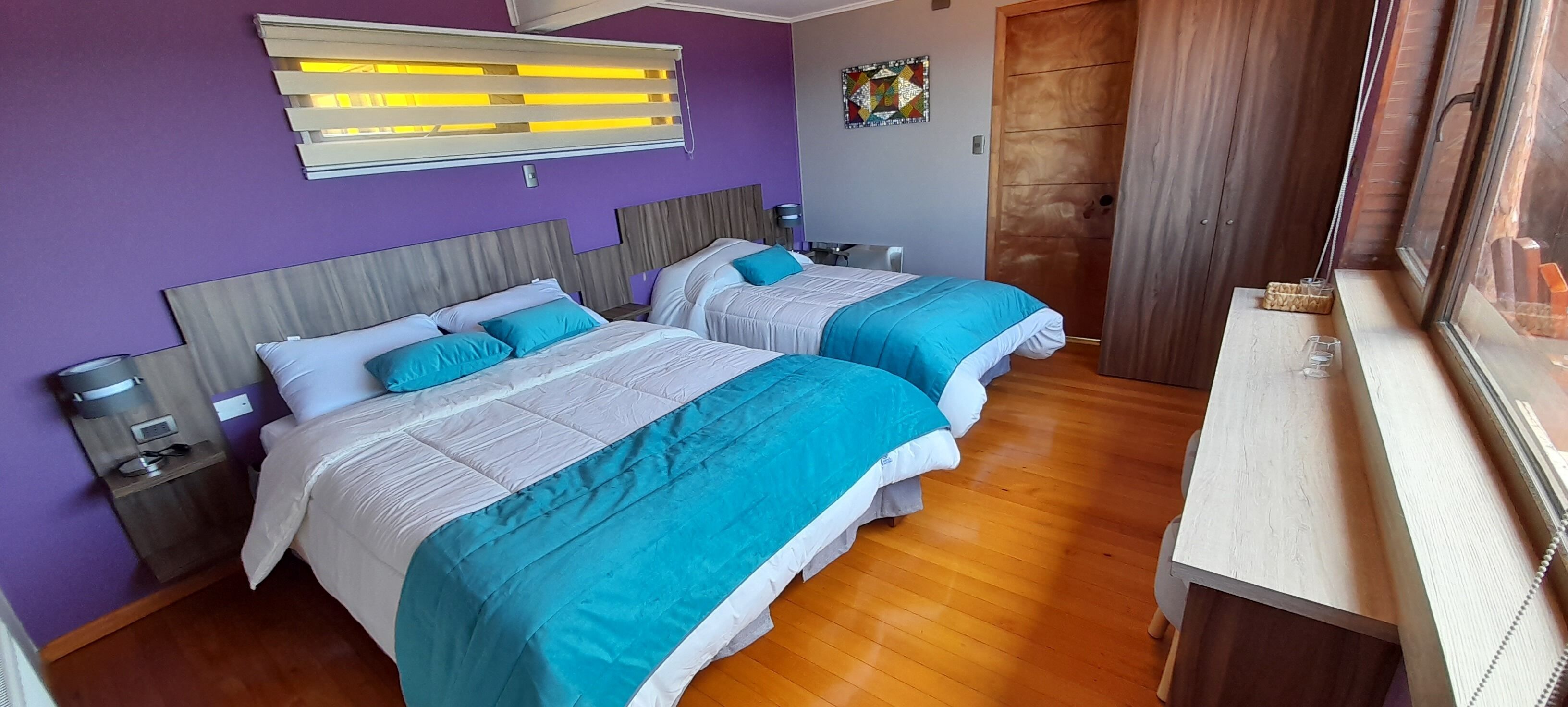 Une chambre d’hôtel avec deux lits, un plancher en bois, une fenêtre avec des stores, une porte et une petite table avec une lampe.