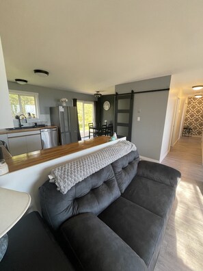 A modern living room with a grey sofa, a wooden coffee table, and a kitchen in the background. - Sunrise Mountain view Skidegate full house, sleeps 8 Haida Gwaii central private (Skidegate)