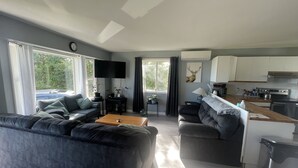 A modern living room with a sectional sofa, a coffee table, and a flat-screen TV. There is a kitchen area with a stove, sink, and cabinets. Large windows provide natural light and a view of the outdoors.