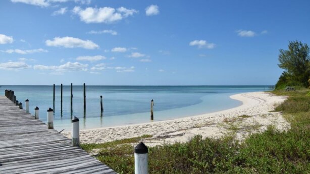 A wooden boardwalk leading to a sandy beach with clear blue water and a few wooden posts.
