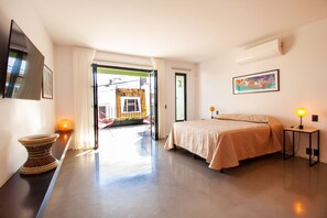 A modern hotel room with a bed, bedside table, and a view through an open door to the outside.