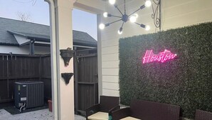 Un patio al aire libre con mobiliario de mimbre, un letrero de neón con la palabra 'Houston' y una pared decorativa.
