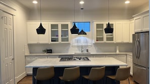 Una cocina moderna con armarios blancos, una isla central con taburetes de bar, iluminación colgante y un refrigerador de acero inoxidable.