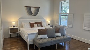 Un dormitorio con cama, mesitas de noche, un banco, una lámpara de pared y una ventana con contraventanas.