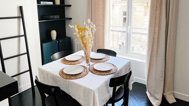 A dining area with a table set for four, a vase with flowers, and a window with curtains.