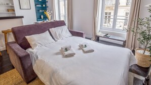 A neatly made bed with white linens, a purple armchair, and a small table with a chessboard.