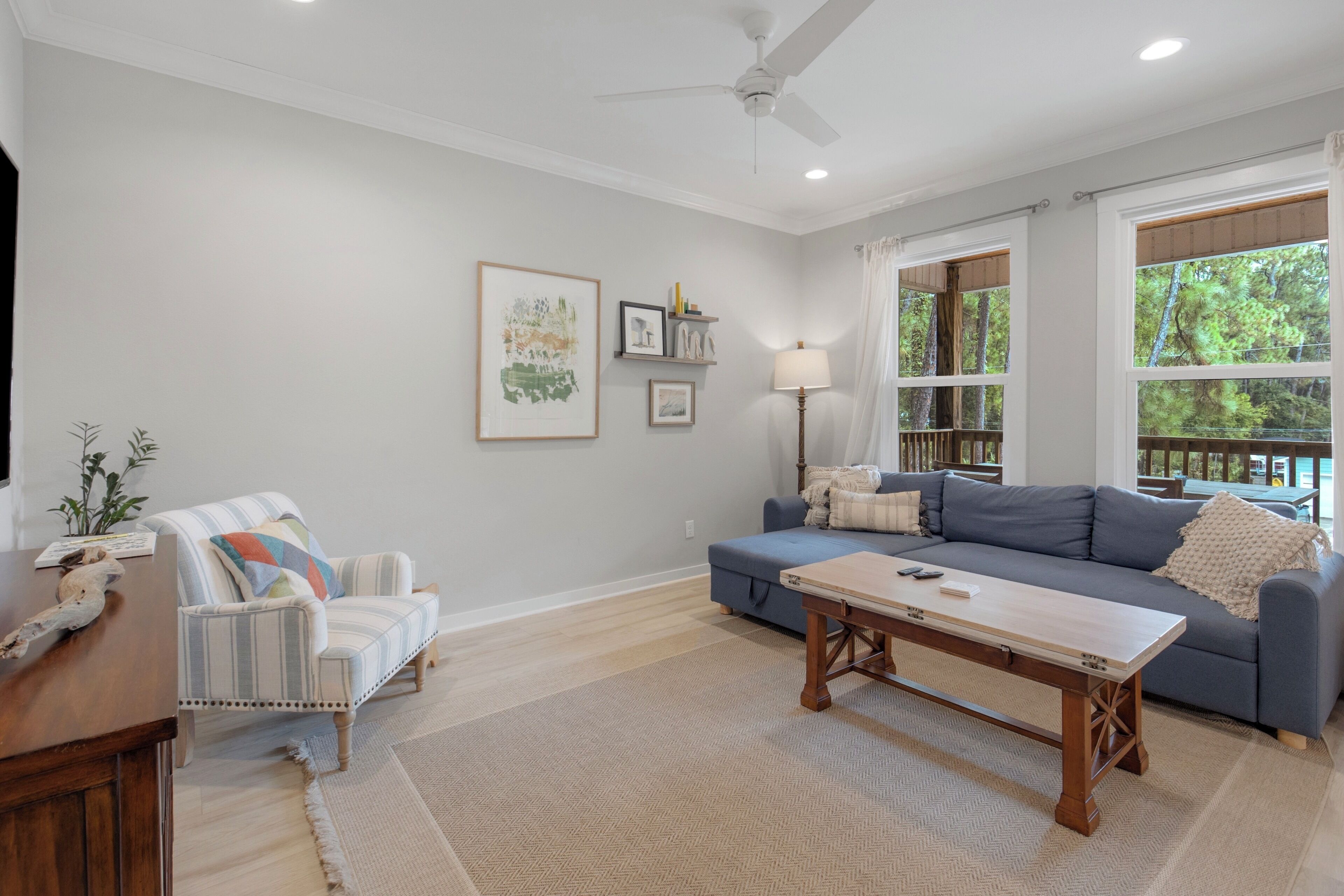 Un salon comprenant un canapé, un fauteuil et une table basse. Les murs sont ornés de tableaux encadrés et un ventilateur de plafond est installé au centre.