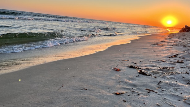 Uma praia ao pôr do sol com ondas, areia e destroços espalhados.