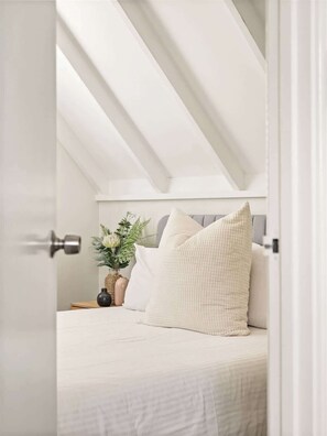 A bedroom with a sloped ceiling, a bed with white bedding, a bedside table with a vase of flowers, and a door with a silver handle.