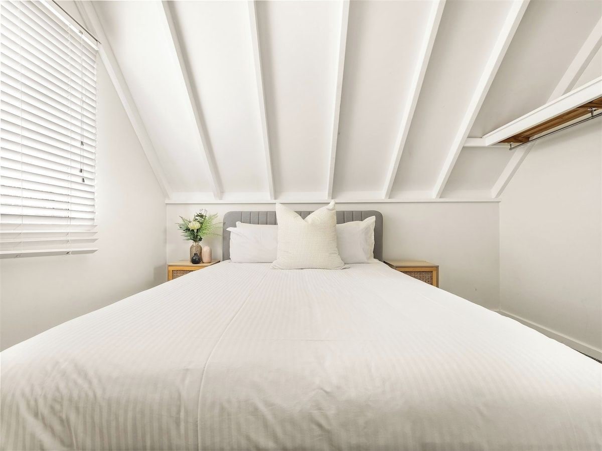 A bedroom with a large bed, a headboard, bedside tables, and a window with blinds.