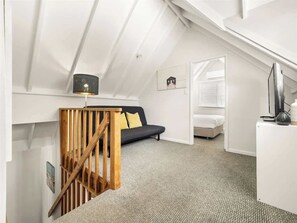 A loft bedroom with a sloped ceiling, a wooden staircase, a black sofa, a white desk, and a TV.