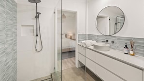 A modern bathroom with a glass-enclosed shower, a round mirror, and a white sink with a soap dispenser.