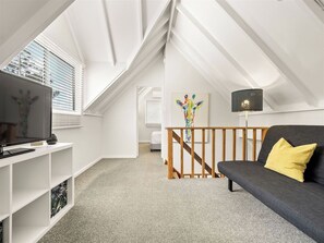 A modern loft bedroom with a sloped ceiling, a grey sofa, a wooden staircase, and a TV on a white console.