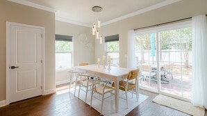 Un comedor con una mesa y sillas blancas, una lámpara de techo y vistas al exterior a través de amplios ventanales.