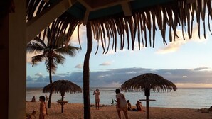 Escena de playa con sombrillas de paja, palmeras y gente relajándose en la arena.