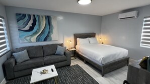 Et moderne hotelværelse med en grå sofa, en seng med hvide sengetøj, et lille bord med en dekorativ genstand og et stort abstrakt maleri på væggen.