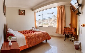 A hotel room with a bed, a small table with a vase of flowers, a desk with a lamp, and a view of buildings through the window. - ALO INKA HOTEL (Cusco)