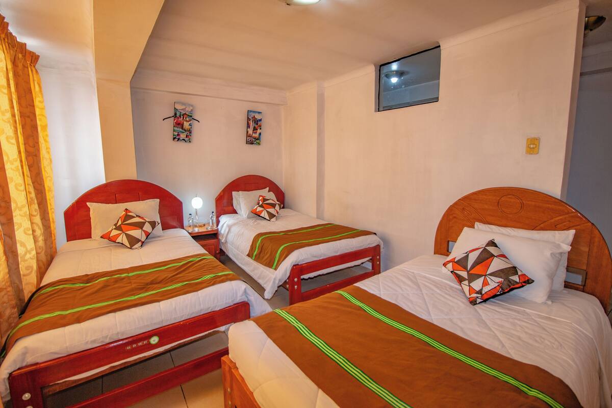 Una habitación con tres camas de madera, cada una con cabecera roja y ropa de cama blanca. Hay dos obras de arte colgadas en la pared y un televisor pequeño.