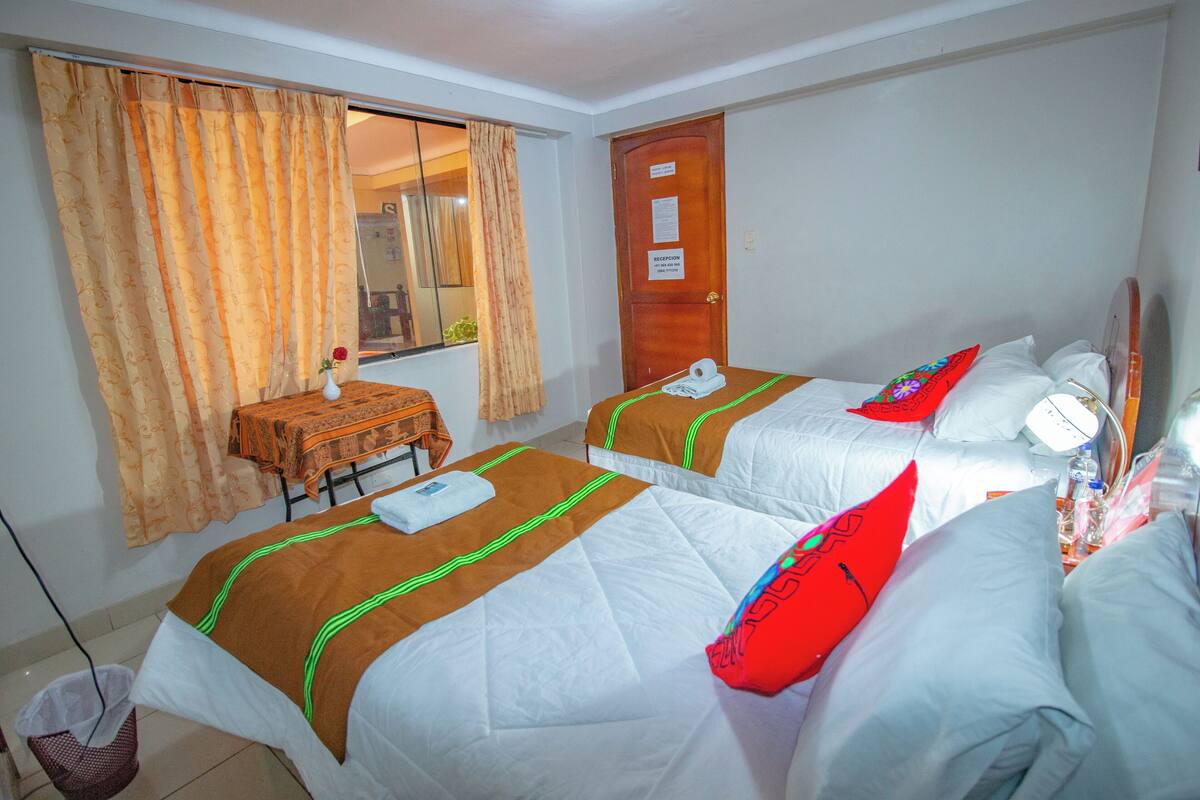 Una habitación de hotel con dos camas, una mesita con un florero y una ventana con cortinas.