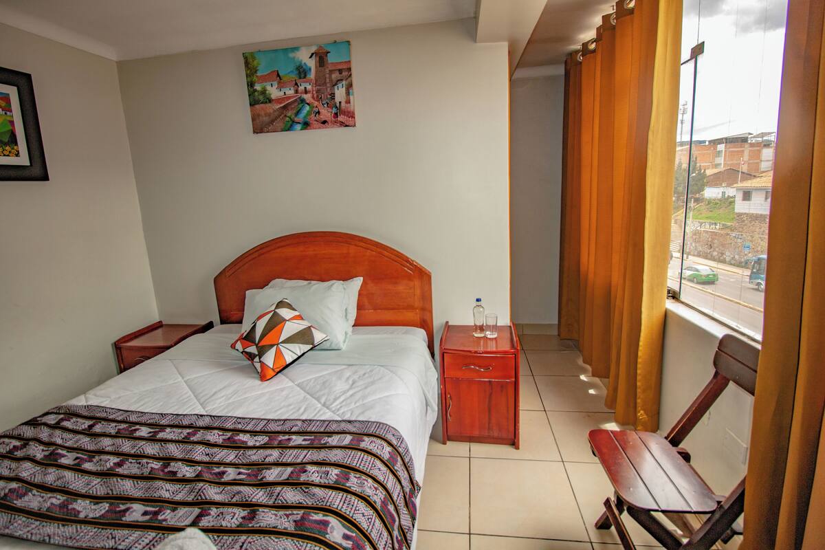 Un dormitorio con cama, mesitas de noche, una silla de madera, ventana con cortinas y un cuadro en la pared.