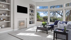 Un salón moderno con televisor de pantalla plana, chimenea y un ventanal con vistas a árboles y una valla.