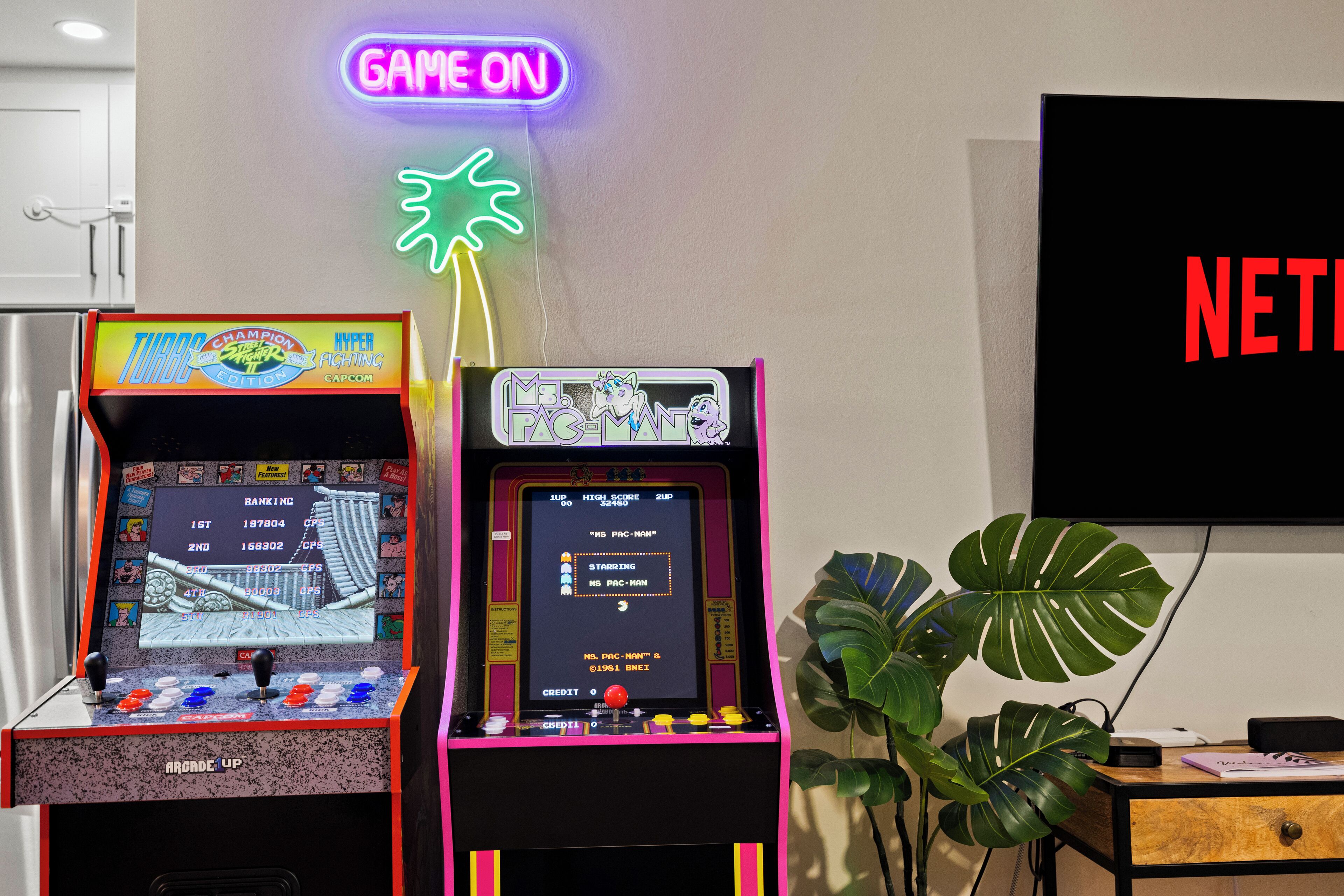Two arcade machines, a neon sign, and a plant in a room.