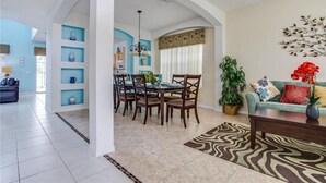 Uma sala ampla com uma mesa de jantar, cadeiras, sofá e mesa de centro. Há uma planta grande e um objeto decorativo na parede.