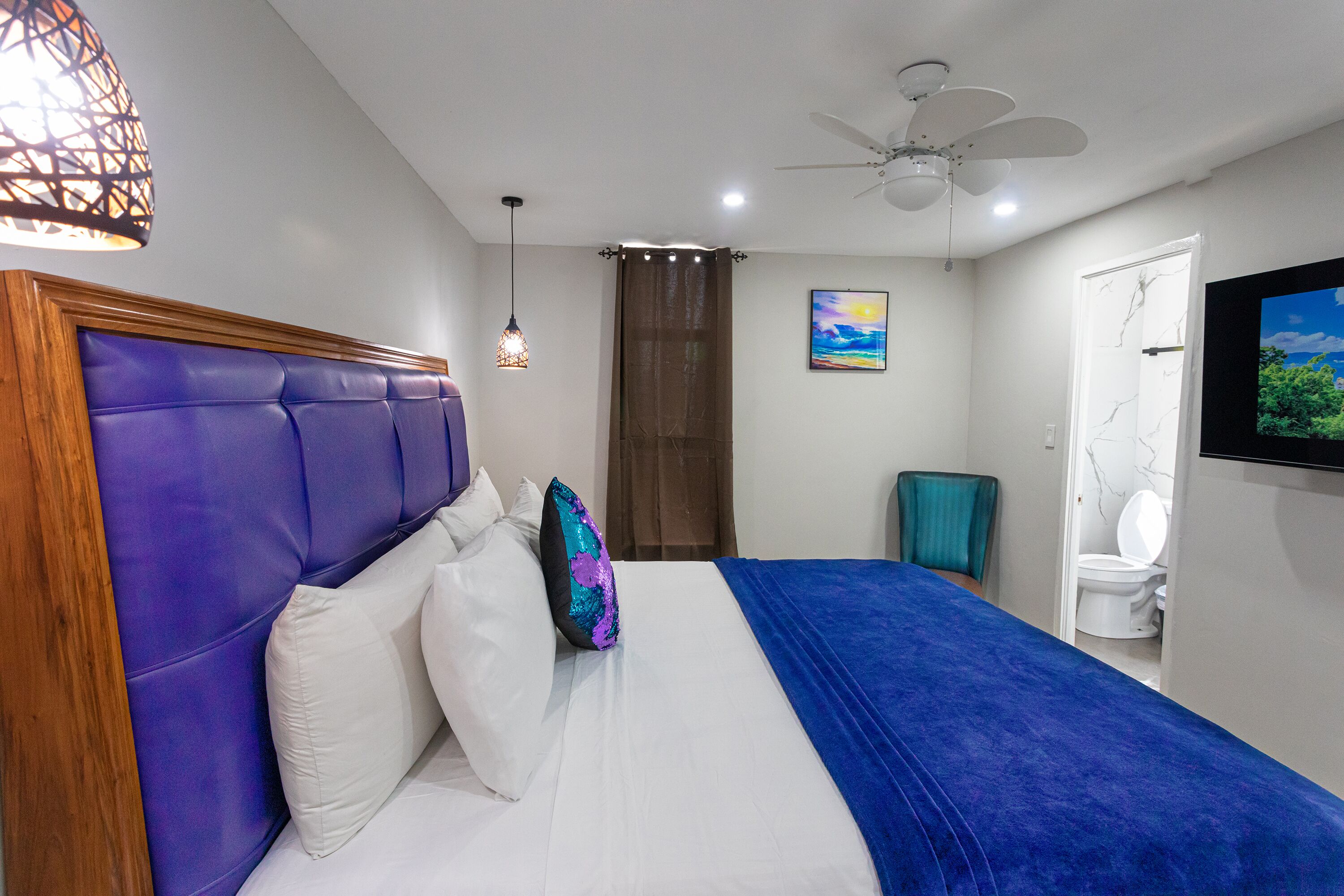 Un dormitorio con una cama grande, un ventilador de techo, un televisor montado en la pared y un baño visible a través de una puerta abierta.