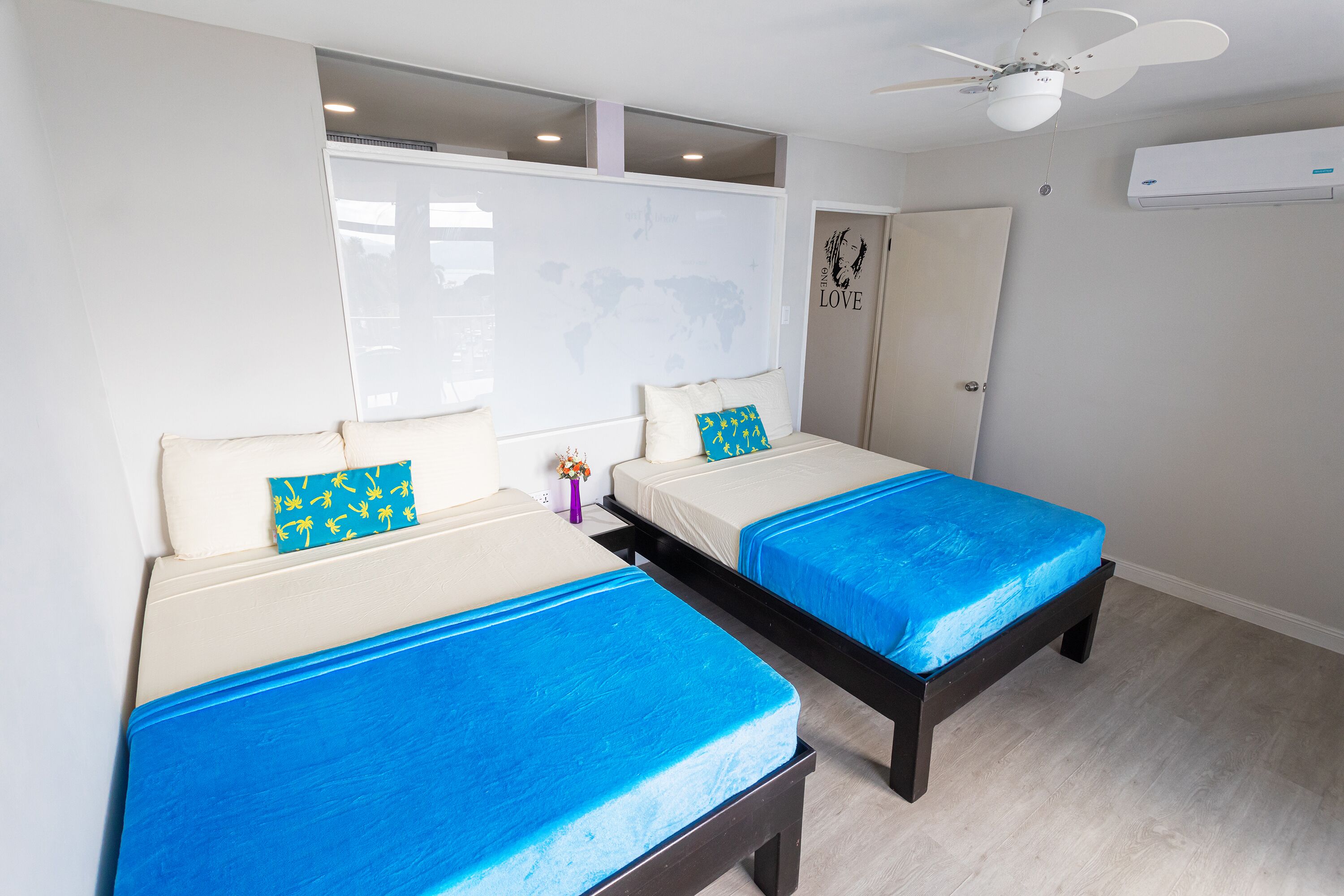A hotel room with two beds, a ceiling fan, a world map on the wall, and an air conditioning unit.