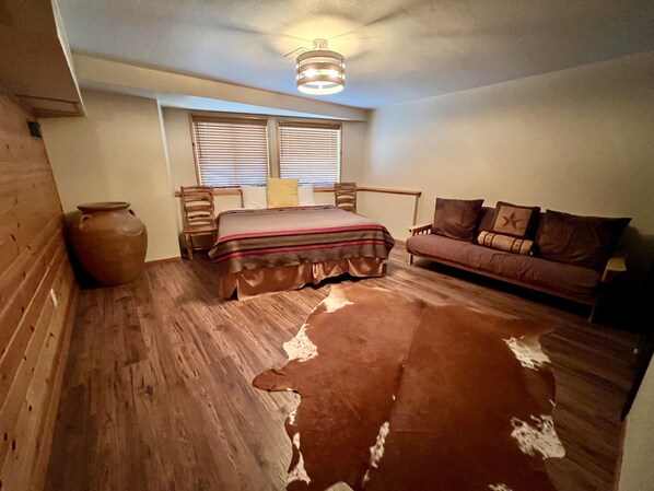 Une chambre avec un plancher en bois, un lit recouvert d’une courtepointe à rayures, un canapé en cuir marron, une chaise en bois et un luminaire suspendu au plafond.