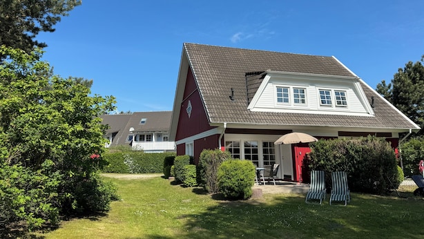 A two-story house with a sloped roof, a front porch, and a small garden area with a lawn and shrubs. - Cozy vacation home for 4-6 persons with garden in Trassenheide (Trassenheide)