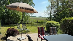 An outdoor patio with a table and chairs, an umbrella, and a view of a grassy field and trees. - Cozy vacation home for 4-6 persons with garden in Trassenheide (Trassenheide)