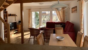 A cozy living room with a wooden staircase, a red sofa, wicker chairs, a dining table with a bowl, and large windows with curtains. - Cozy vacation home for 4-6 persons with garden in Trassenheide (Trassenheide)