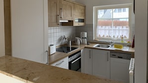 A modern kitchen with white cabinets, a built-in oven, a coffee maker, and a window with a view of a fence and trees. - Cozy vacation home for 4-6 persons with garden in Trassenheide (Trassenheide)
